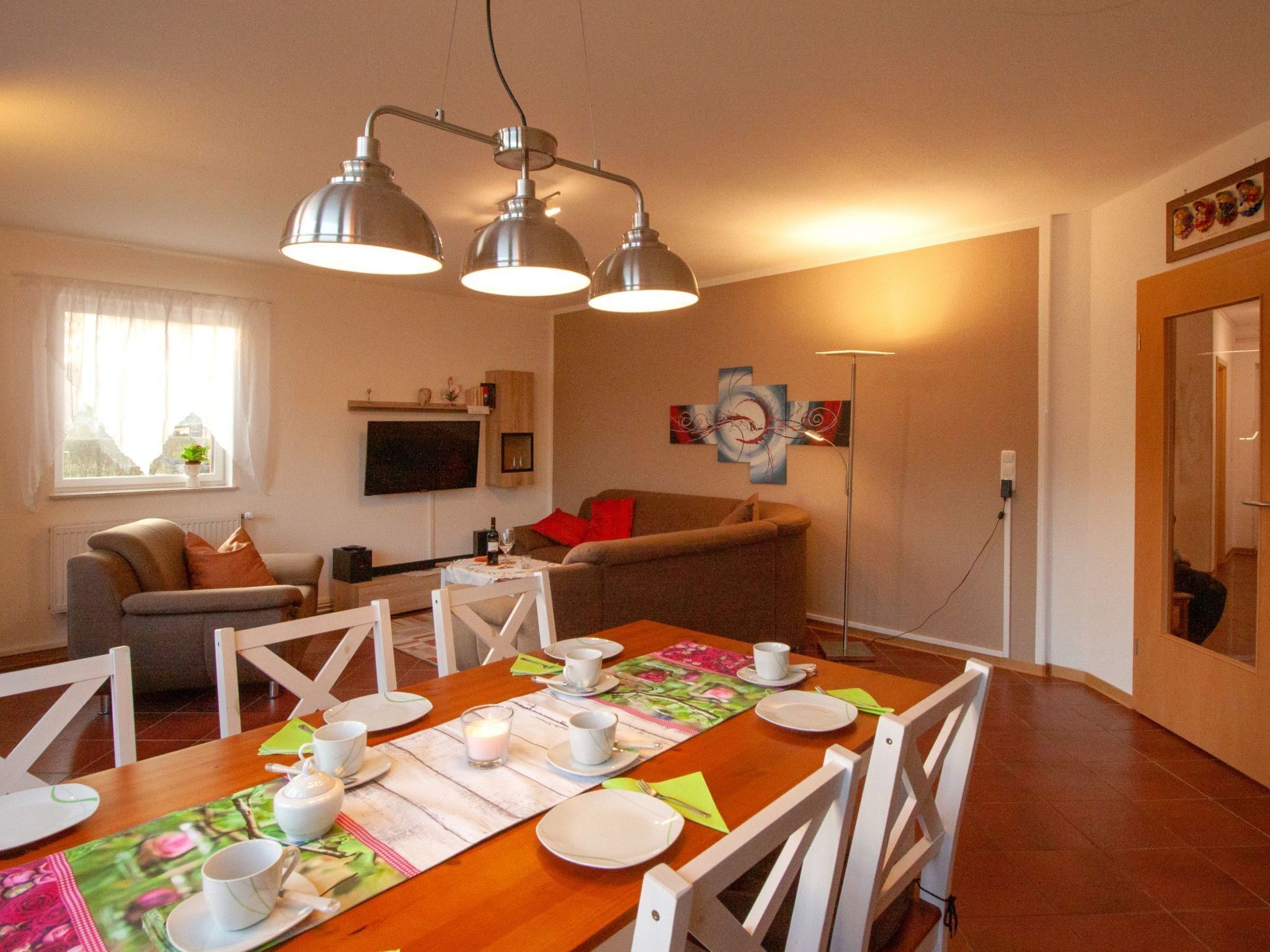 Un coin repas avec une table en bois dressée pour quatre personnes, entourée de chaises blanches. On aperçoit un canapé et un téléviseur au fond.