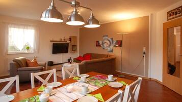 Un coin repas avec une table en bois dressée pour quatre personnes, entourée de chaises blanches. On aperçoit un canapé et un téléviseur au fond.