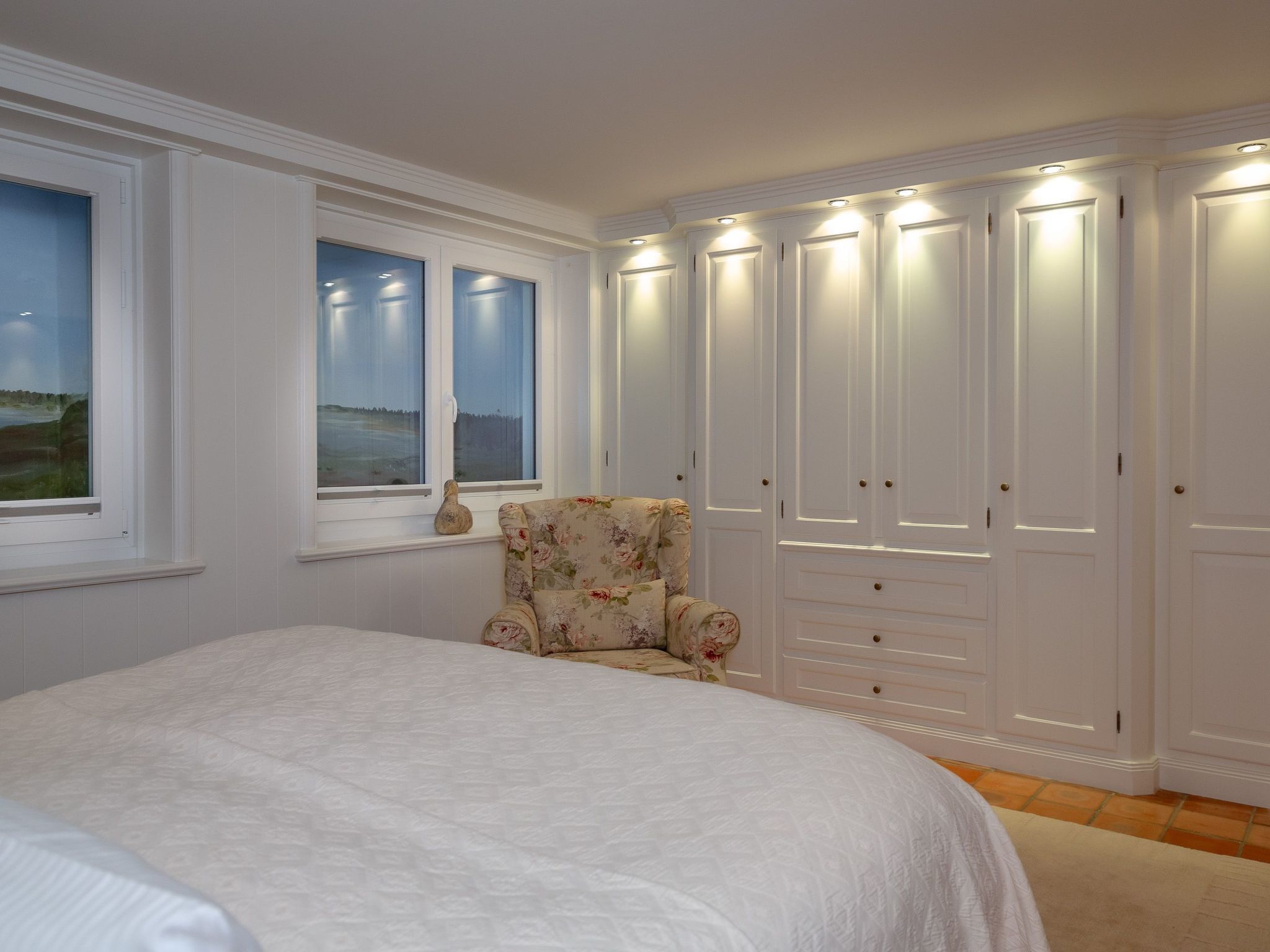 Une chambre avec un grand lit, un fauteuil à motifs floraux, une armoire aux portes éclairées et une fenêtre donnant sur un paysage.