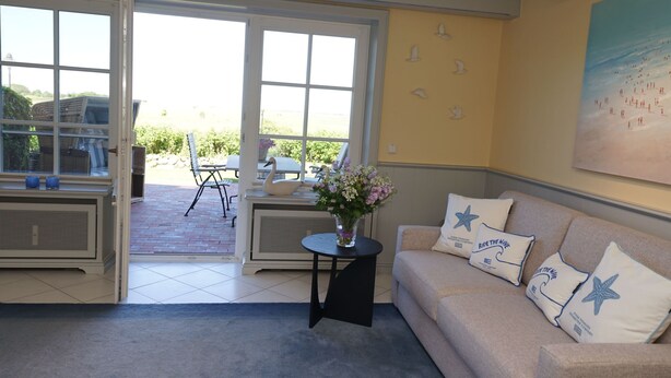 Une pièce avec un canapé beige, une petite table sur laquelle est posé un vase de fleurs et une grande fenêtre donnant sur l’extérieur.