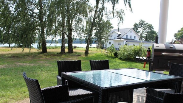 Un patio al aire libre con una mesa de vidrio, sillas de mimbre, una parrilla y vistas a un lago y una casa al fondo.