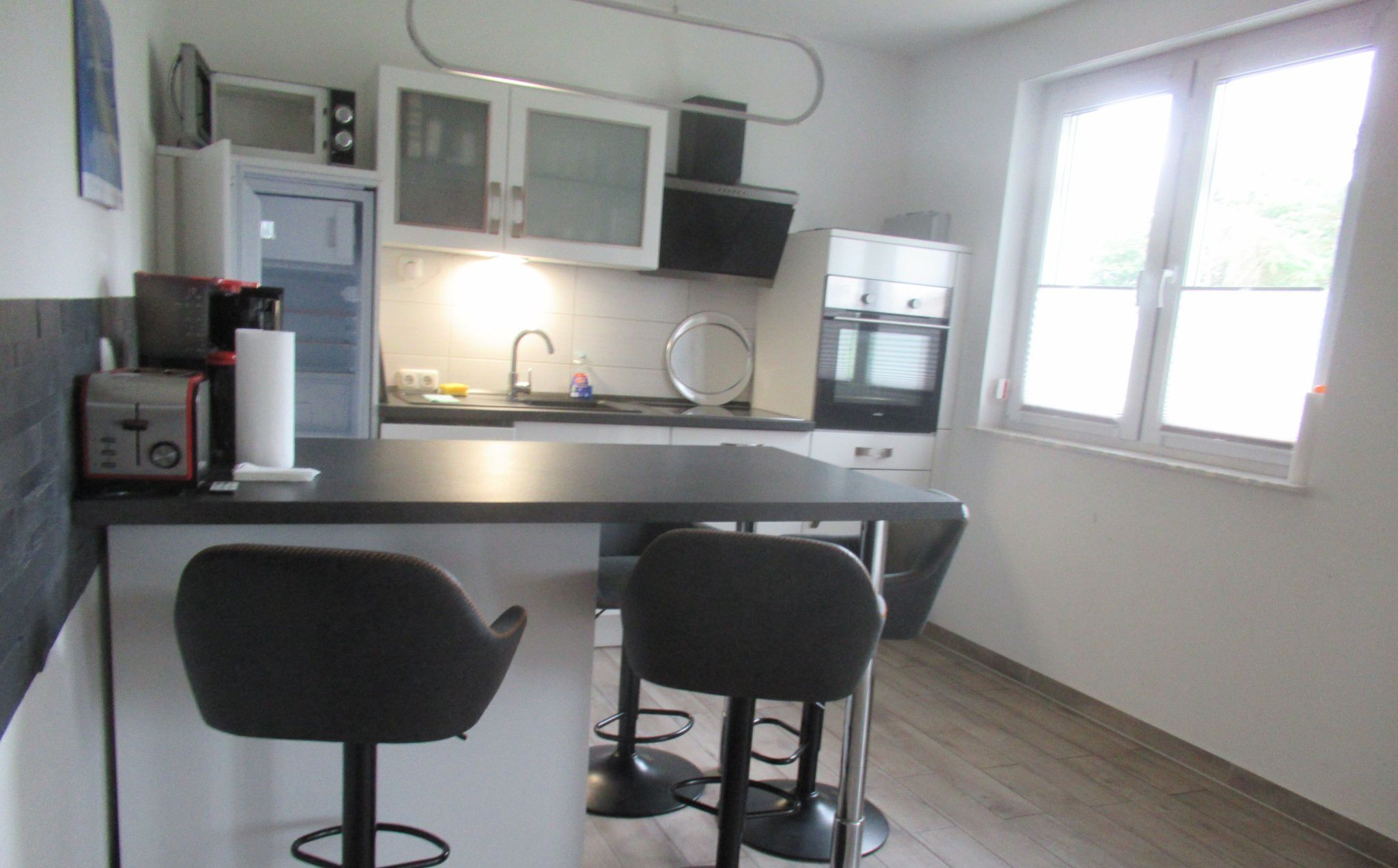 A compact kitchen with a central island, two bar stools, a microwave, a toaster, and a window with blinds.
