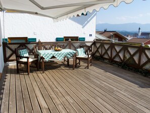 Eine Holzterrasse mit Tisch und zwei Stühlen, eine weiße Wand mit Fenster, und im Hintergrund Häuser und Berge.