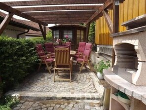 Eine überdachte Terrasse mit Steinboden, Holzmöbeln und einem kleinen Tisch mit Stühlen.