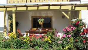 En gul byggnad med en överbyggd veranda, ett matbord med stolar och hängande blomkorgar.