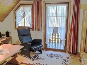 Una habitación con una butaca de cuero negro, una mesa con papeles, un radiador, un tapete estampado y una ventana con cortinas.