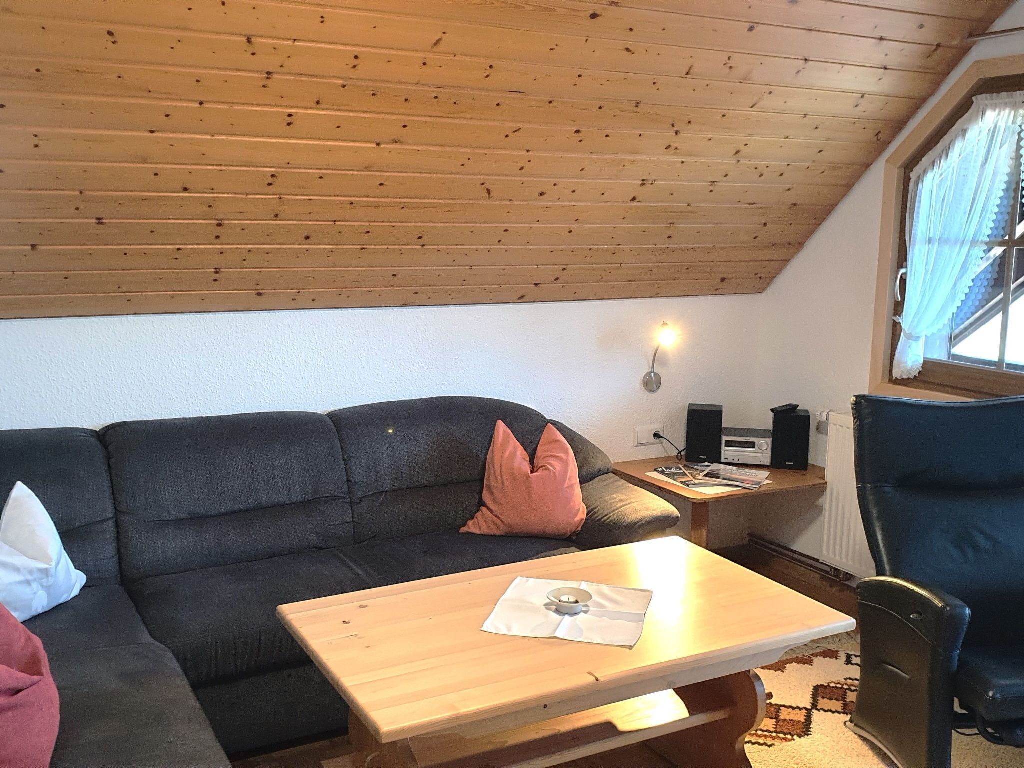 A cozy room with a wooden ceiling, a black sofa with cushions, a small wooden table, and a chair.