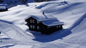 Una cabaña de madera cubierta de nieve con techo inclinado, ubicada en la ladera de una montaña nevada.