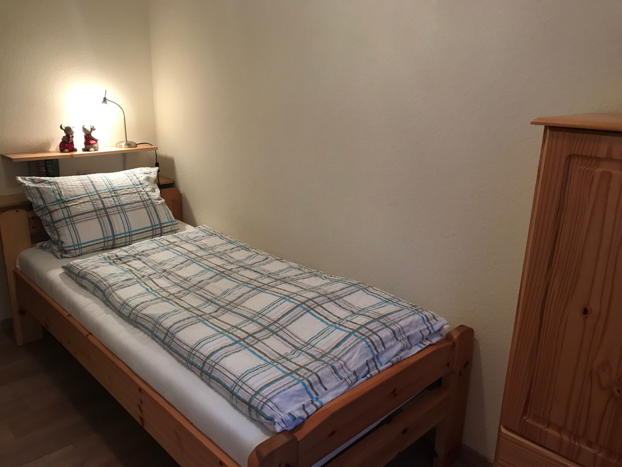 A small, neatly made bed with a plaid bedspread and pillows, a bedside lamp, a small shelf with figurines, and a wooden cabinet.