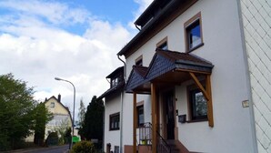Ein zweistöckiges Gebäude mit weißer Fassade und braunen Verzierungen, inklusive einer überdachten Veranda und einer Treppe zum Eingang.