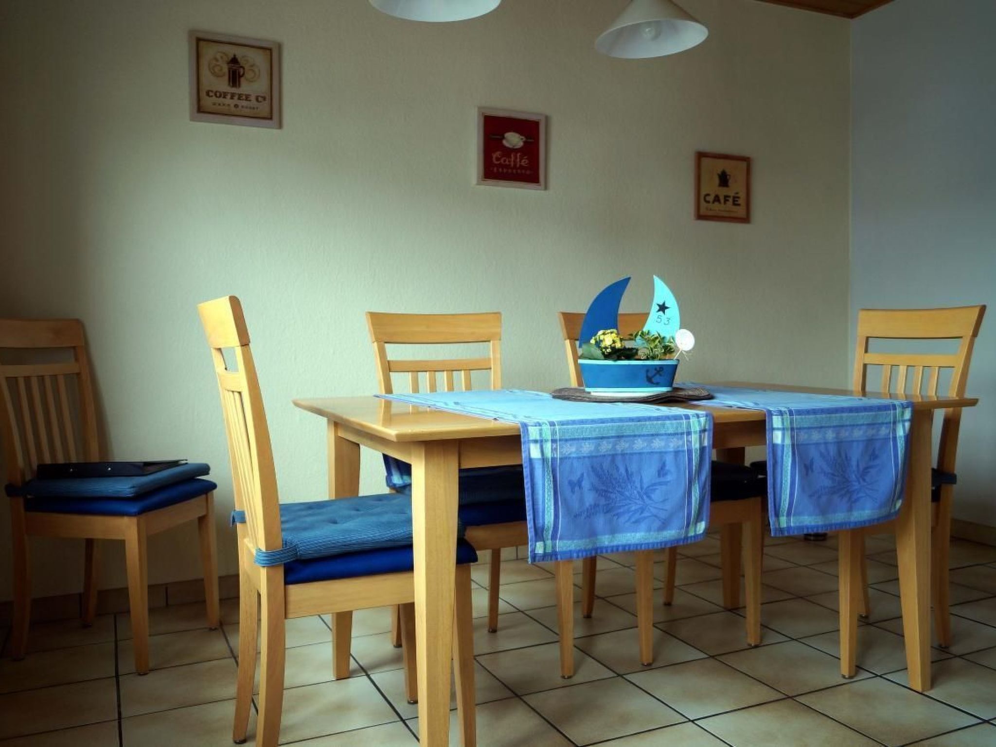 Un coin repas avec une table et des chaises en bois, une nappe bleue et un bol décoratif sur la table, orné d’une image de voilier.
