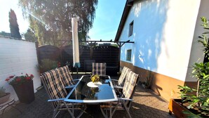 Eine Terrasse mit einem Glastisch und vier Stühlen, umgeben von Topfpflanzen und einem Holzzaun.