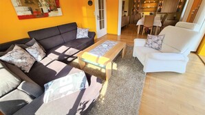 Ein Wohnzimmer mit einer Couch, einem Sessel und einem Couchtisch, einem Gemälde an der Wand und einer Tür, die in eine Küche führt.