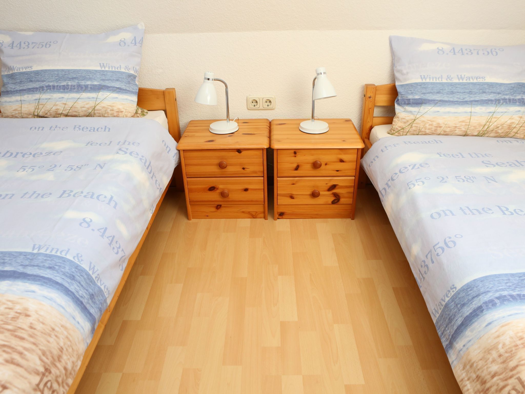 Camera da letto con due comodini in legno, ognuno con una lampada, e un letto con copripiumino a tema spiaggia.
