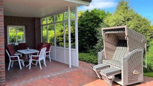 A covered patio with a brick wall, a table with chairs, and a wicker lounge chair. - Strandnah Ilse Hans by Interhome (Friederikensiel)