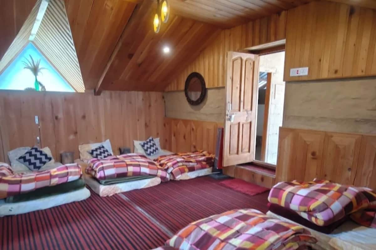 A cozy room with wooden walls and sloped ceilings, featuring multiple beds with colorful bedding and a small window with a view of palm trees.