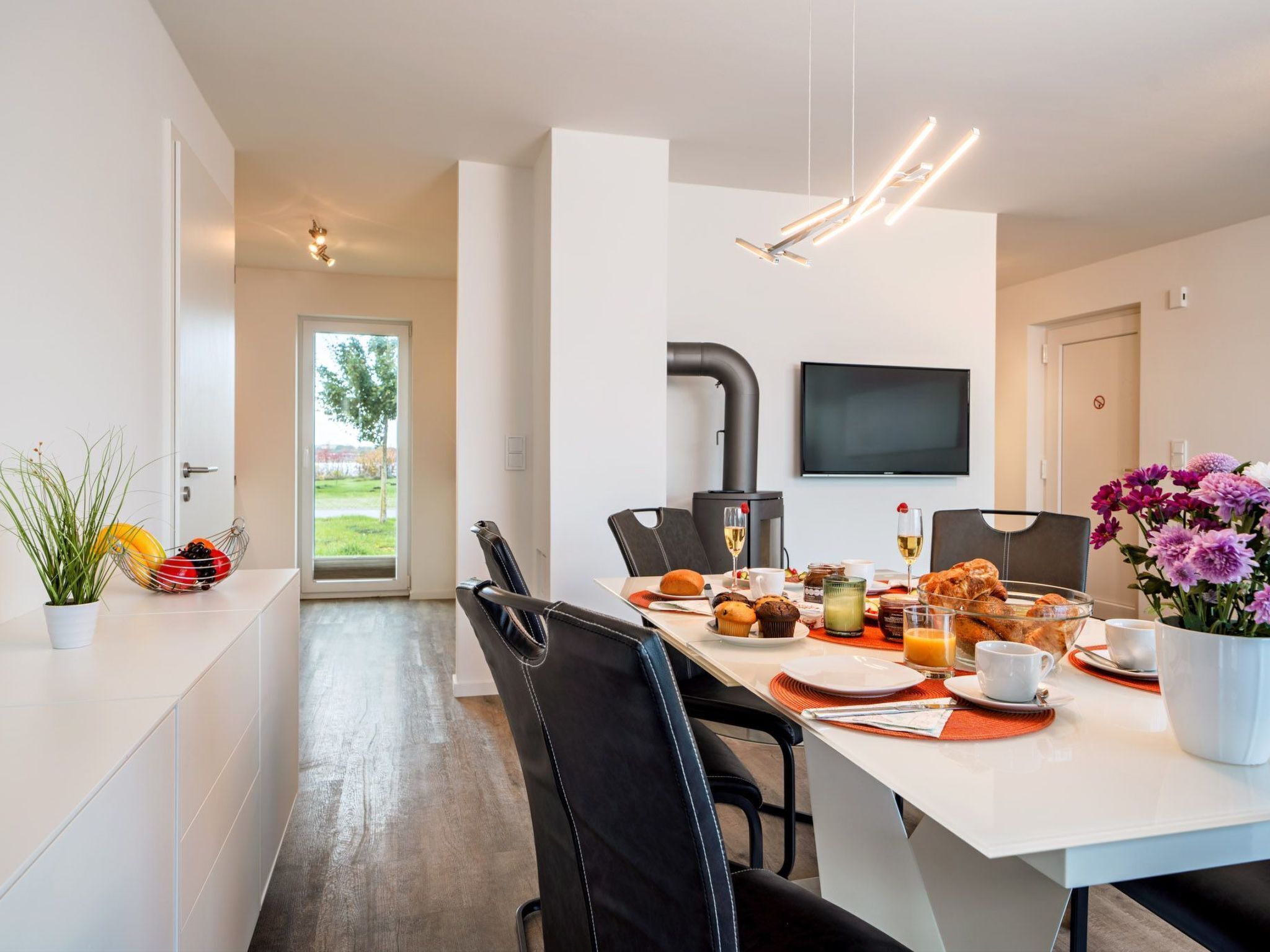 A modern dining area with a white table set for breakfast, featuring pastries, fruits, and beverages.