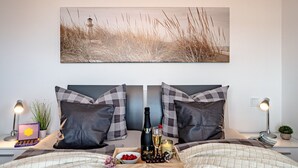 Una cama bien hecha con un juego de almohadas a cuadros, una bandeja con aperitivos y una botella de champán, y un cuadro enmarcado sobre la cama.