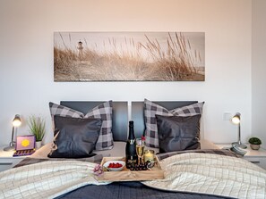 A neatly made bed with a checkered pillow set, a tray with snacks and a bottle of champagne, and a framed picture above the bed. - Schleivilla Lootsen Hus by Seeblick Ferien ORO by Interhome (Hasselberg)