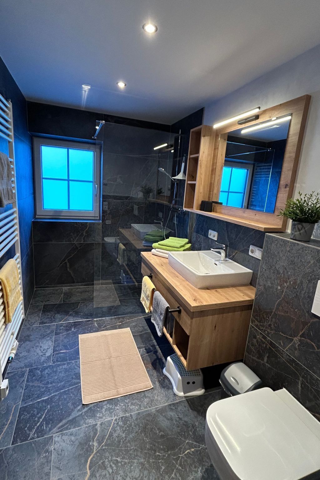 Une salle de bain moderne avec un meuble-lavabo en bois, un grand miroir, une douche intégrée et un sol en marbre foncé.