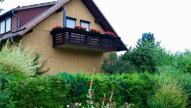 Een tweelaags huis met een bakstenen gevel, een balkon met rode bloemen en een tuin met diverse planten en bloemen.