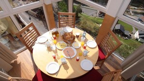 Une table haute, dressée pour le petit-déjeuner, entourée de chaises en bois avec des coussins rouges, dans une pièce dotée de grandes fenêtres donnant sur l’extérieur.