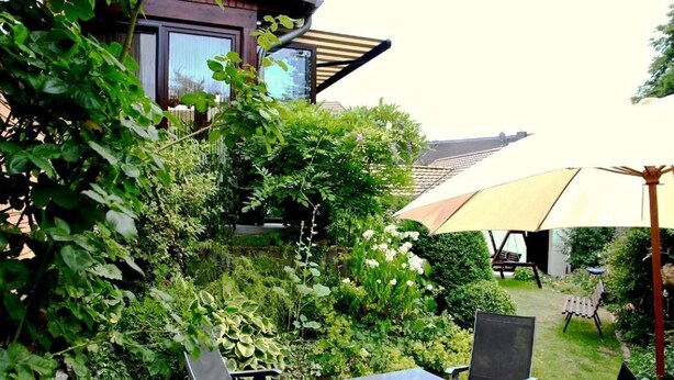 En udendørs patio med et bord og stole, omgivet af frodig vegetation og en stor parasol, der giver skygge.