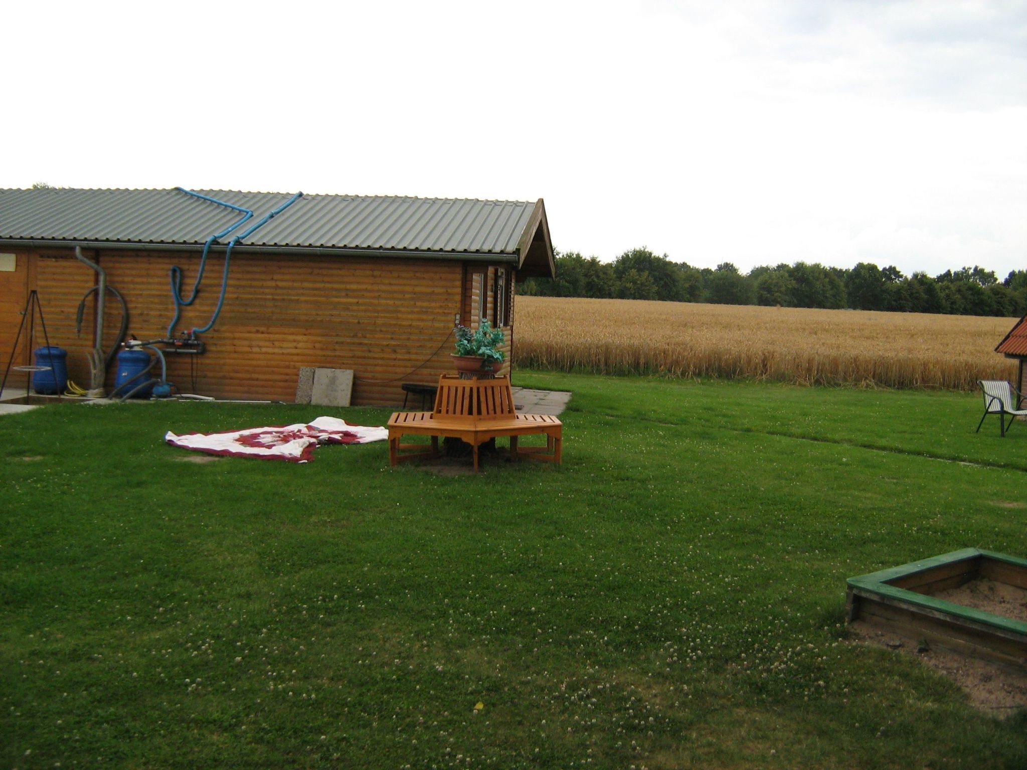 Ein Holzhaus mit Metall-Dach, einem Rasen, einer Holzbank und einem Feld im Hintergrund.