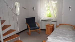 Un dormitorio con cama de madera, una silla, una mesita de noche, un radiador, una ventana con cortinas y una escalera.