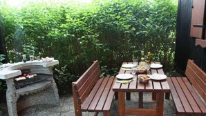 Een buitenterras met een stenen barbecuegrill, houten banken en een tafel gedekt voor twee personen.