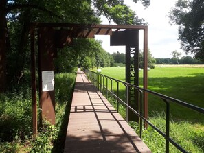 Ein hölzerner Steg mit Metallgeländern, der durch ein grünes Feld mit Bäumen im Hintergrund führt.
