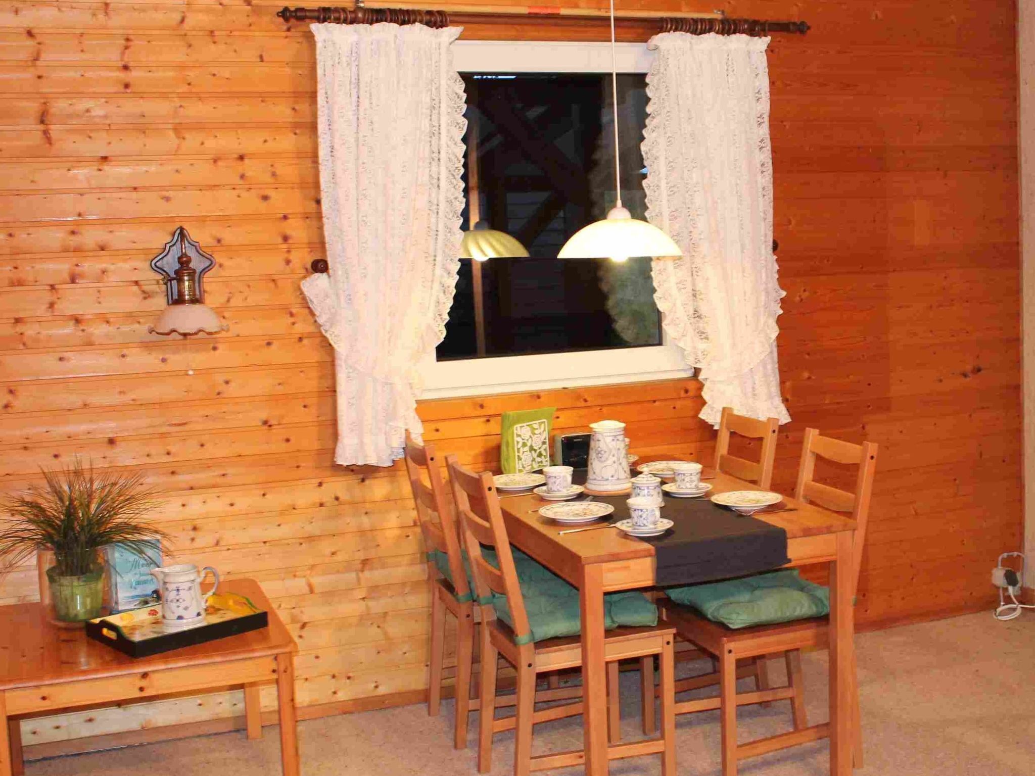 Et interiør af en træhytte med et spisebord dækket til fire personer, omgivet af træstole med grønne hynder. Et vindue med hvide gardiner og en hængende lampe er synligt.