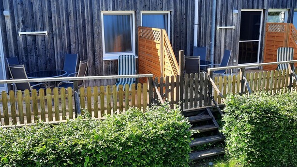 Een houten gebouw met een houten omheining, een kleine trap en een tuin met struiken.