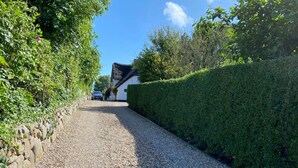 Une allée de gravier mène à une maison à l’extérieur blanc et au toit noir, entourée d’une végétation luxuriante et d’un mur de pierres sur un de ses côtés.