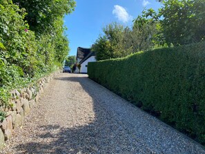 Ein Kiesweg führt zu einem Haus mit weißer Fassade und schwarzem Dach, umgeben von dichter Vegetation und einer Steinmauer auf einer Seite.