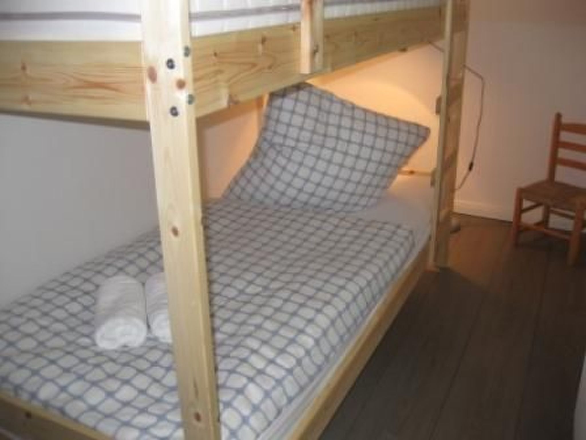 A single bunk bed with a checkered mattress and a pillow, a wooden chair, and a wall socket.