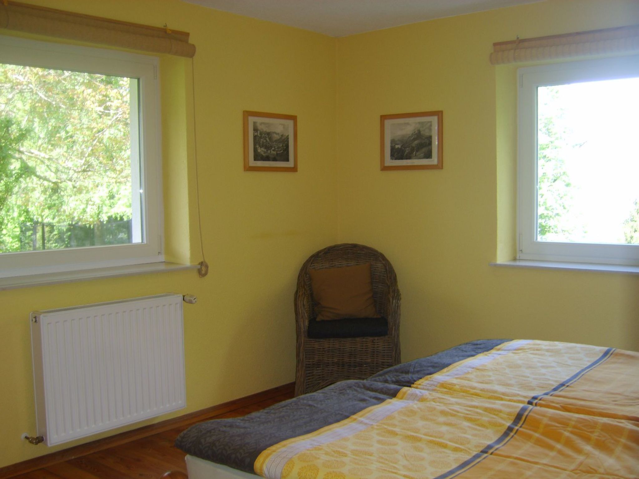 A room with a bed, two windows, a radiator, and two framed pictures on the wall.