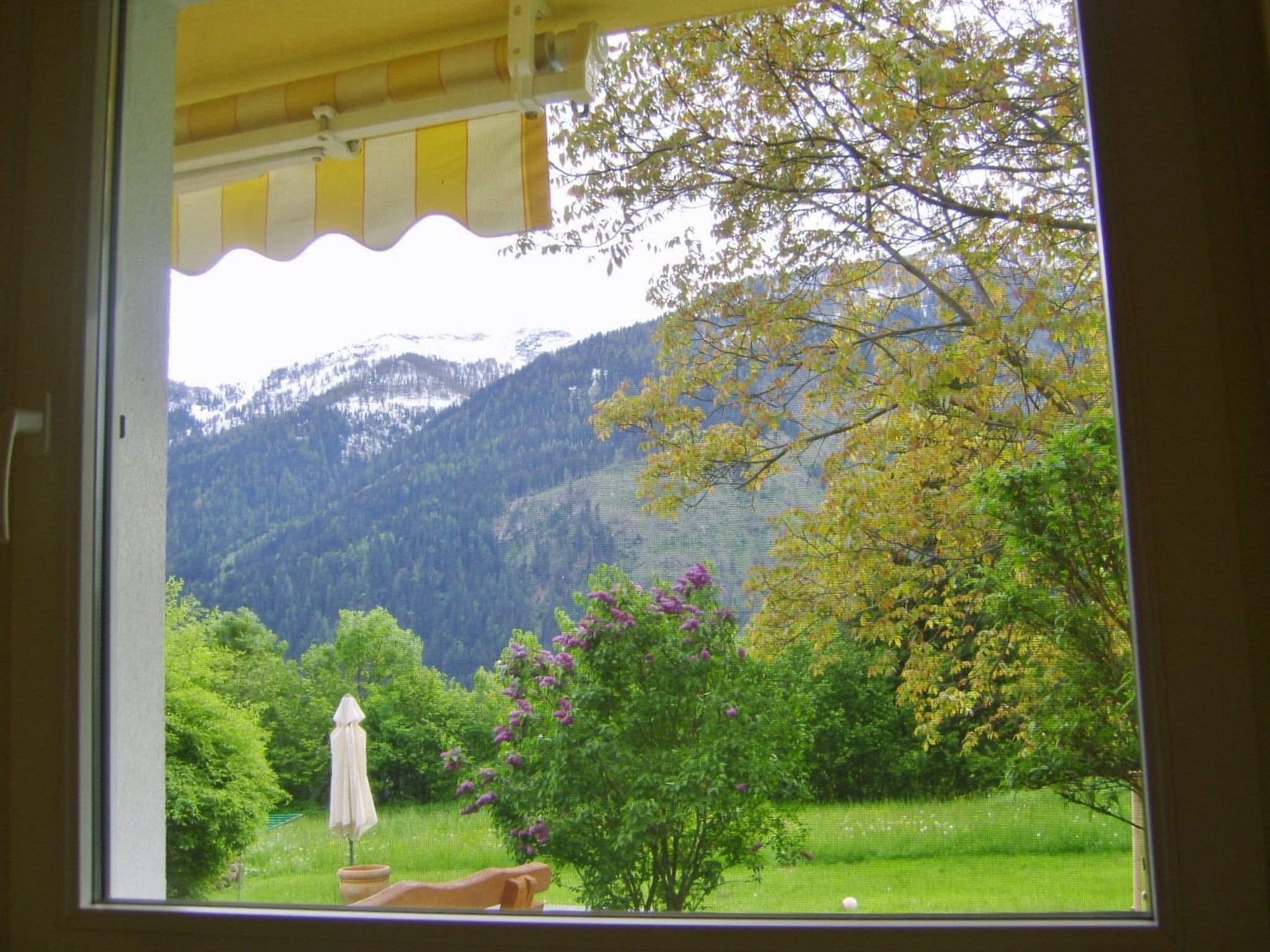 Vista da janela de uma paisagem montanhosa com área gramada e um pátio com guarda-sol.