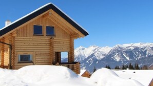 Uma cabana de tronco com telhado em forma de 'A' e duas janelas, rodeada por neve e montanhas.