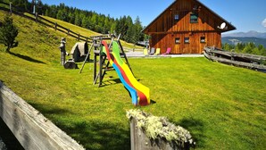 Una cabaña de madera con un tobogán colorido en un área verde, rodeada de árboles y montañas.