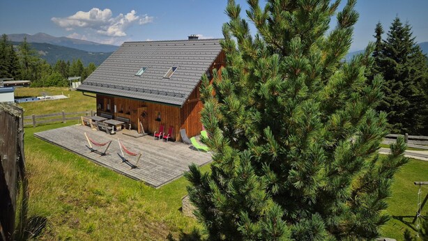 Una cabaña de madera con techo inclinado, una terraza con asientos y un gran pino en primer plano.