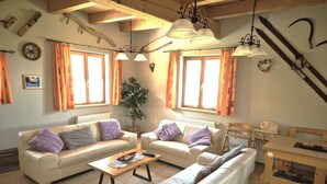 Una sala de estar acogedora con dos sofás, una mesita de centro y una mesa de comedor de madera. Hay cortinas, una planta y un reloj de pared.