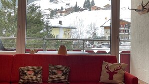 Un divano rosso con cuscini a quadretti e un cuscino a forma di cervo, un vaso e una vista sulle montagne attraverso ampie finestre.