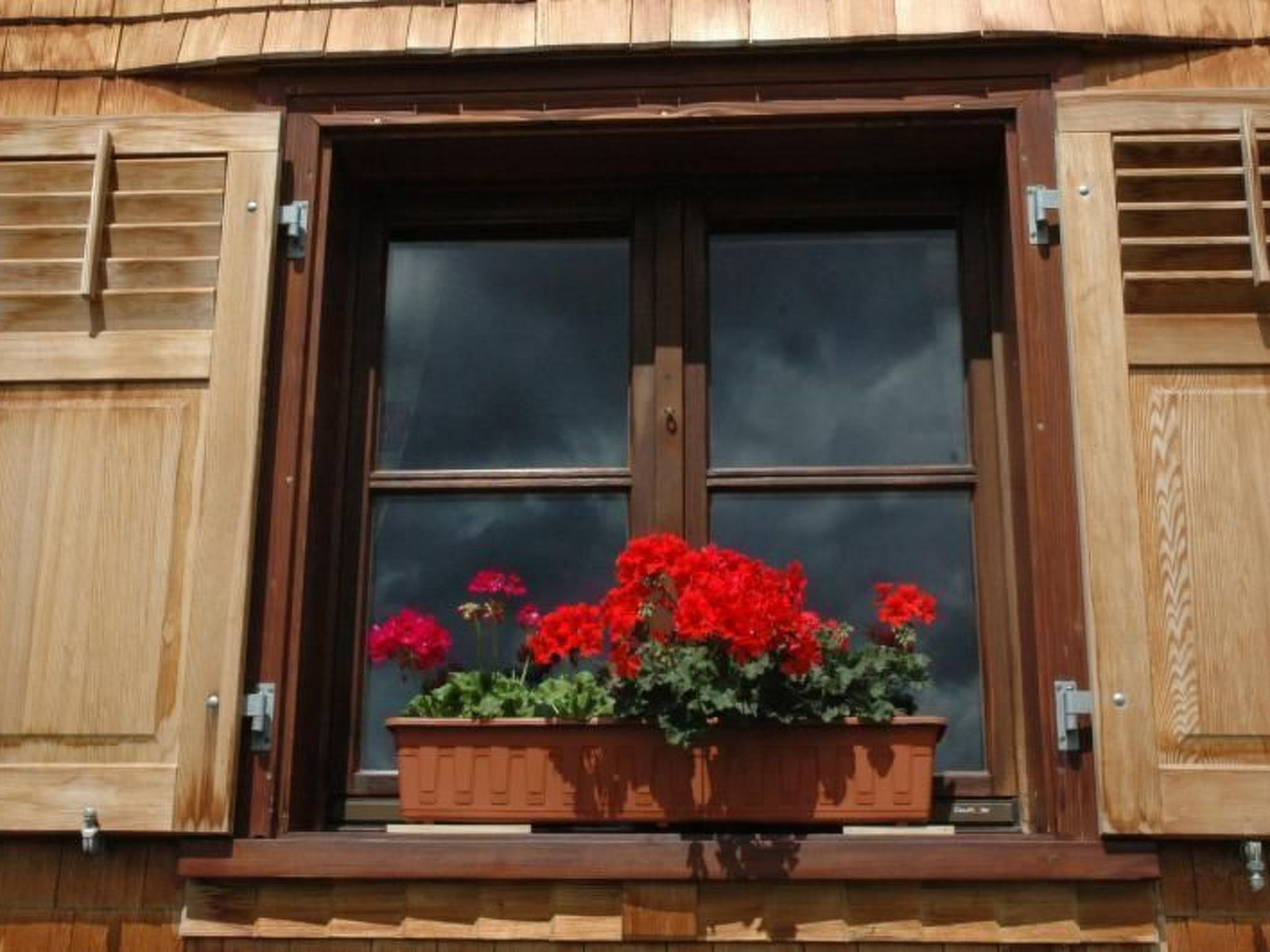 Una ventana con contraventanas, un marco de madera y una maceta con flores rojas.