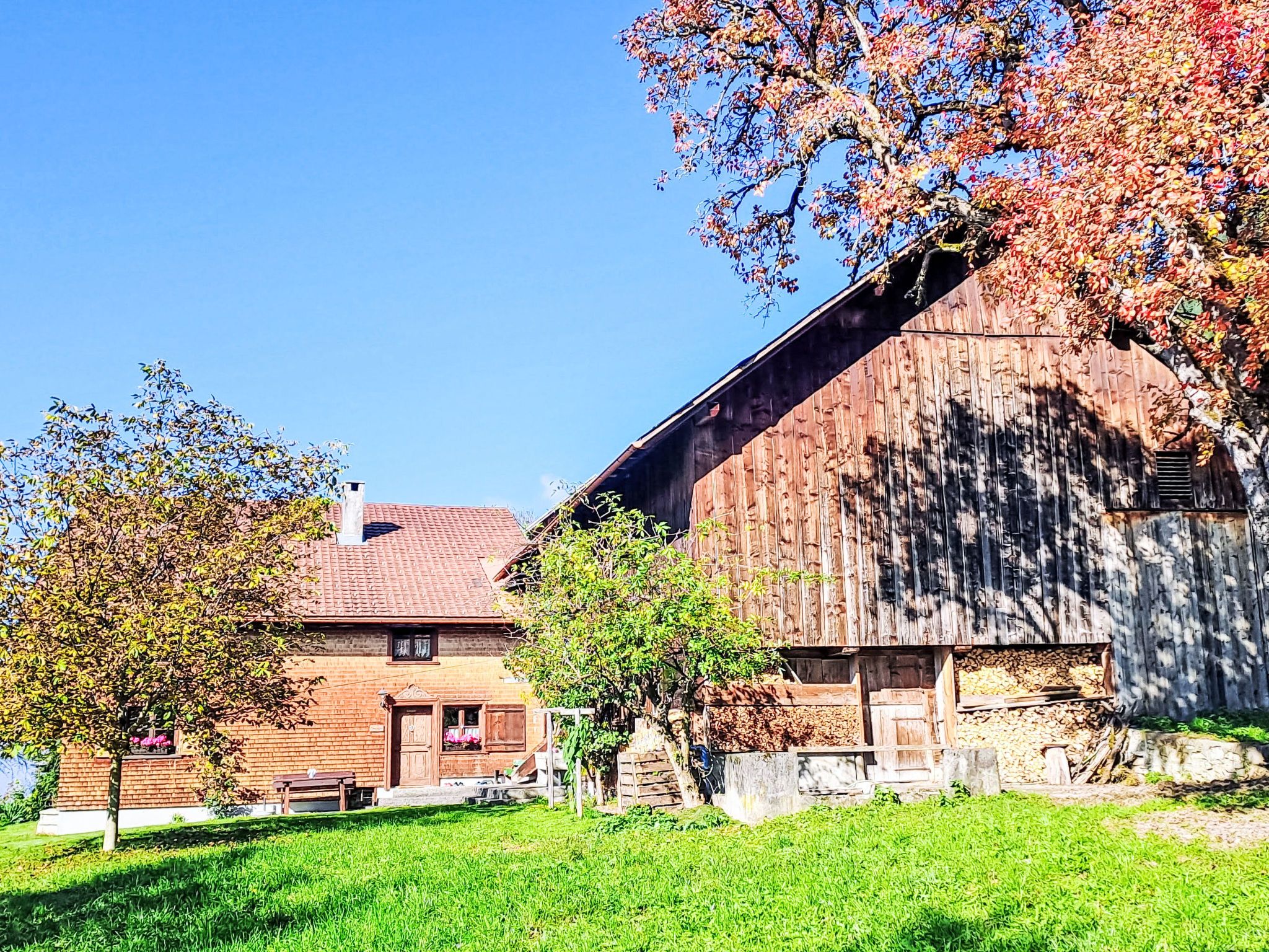 Une grange traditionnelle en bois, dotée d’un toit en pente, entourée d’une pelouse bien entretenue et d’une maison en briques avec un toit en tuiles.