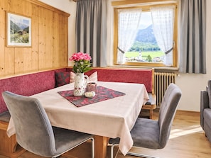Un coin repas avec une table dressée pour deux personnes, un banc agrémenté de coussins, un vase rempli de fleurs et une fenêtre offrant une vue sur un paysage montagneux.