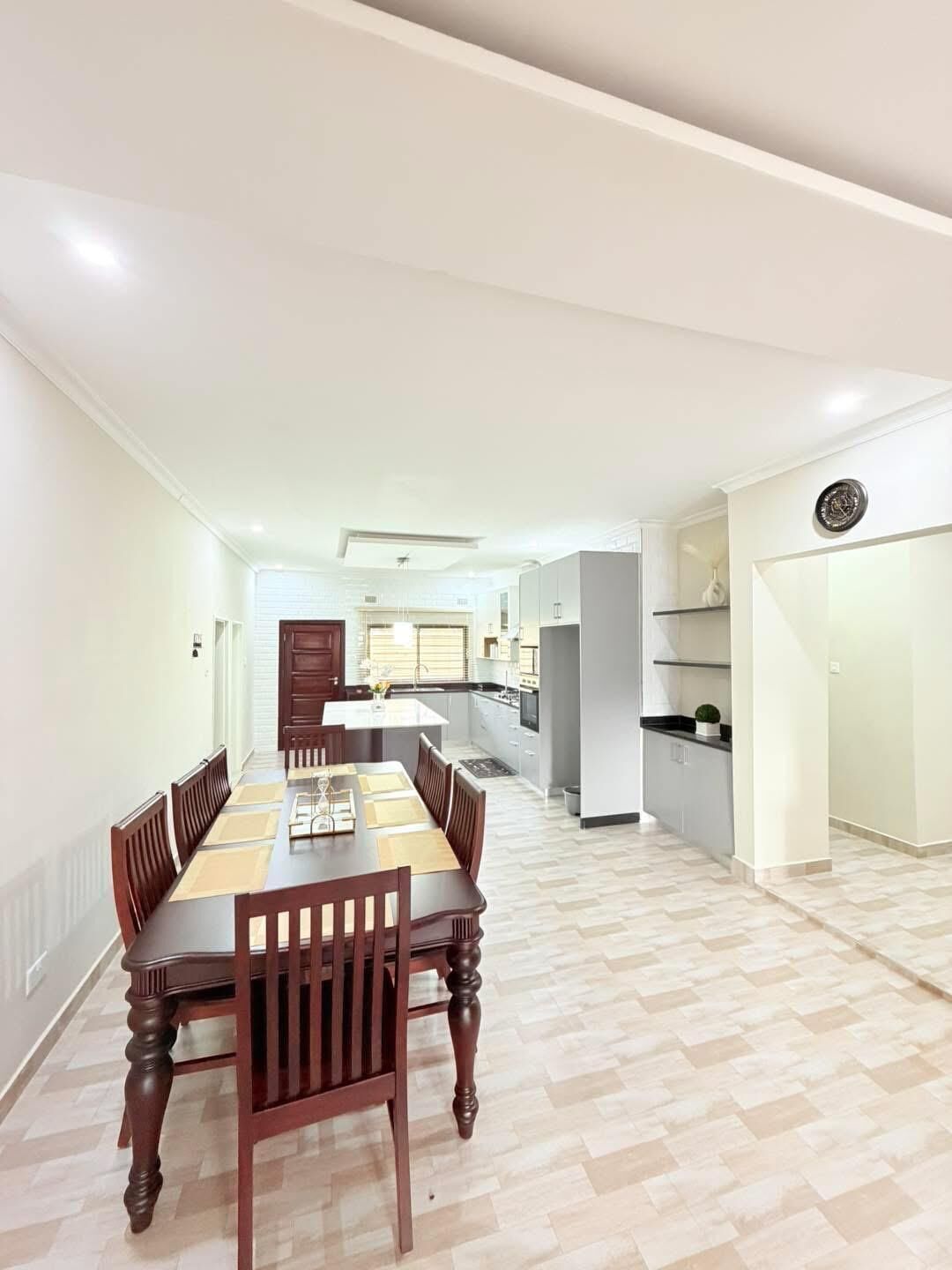 A spacious dining area with a long wooden table and chairs, a kitchen in the background, and a modern interior design.