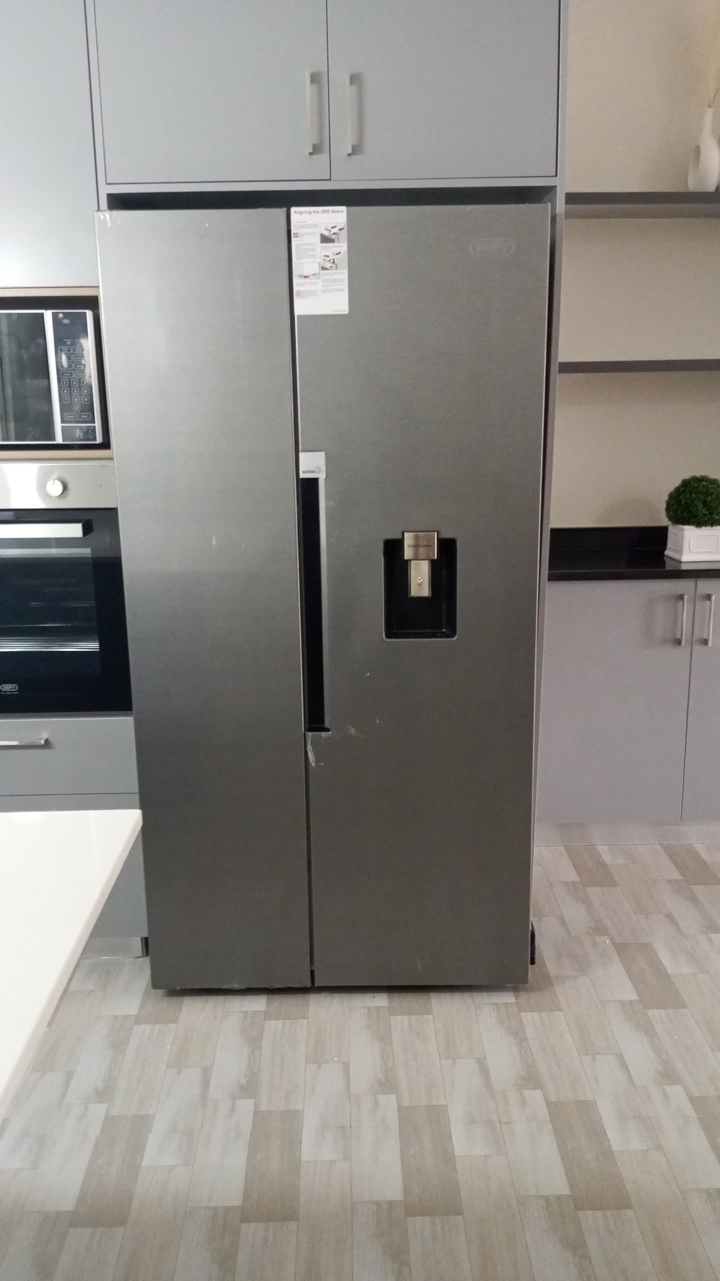 A stainless steel refrigerator with a water dispenser, located in a kitchen with light wood flooring and grey cabinets.