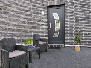 Fachada moderna de un edificio con pared de ladrillo gris oscuro, puerta negra de diseño único y mobiliario exterior con sillas de mimbre y una pequeña mesa.