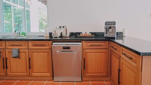A kitchen with wooden cabinets, a dishwasher, a coffee maker, and a window with a view of greenery. - Villa 7 - Luxury Private Pool Villa (Langkawi)